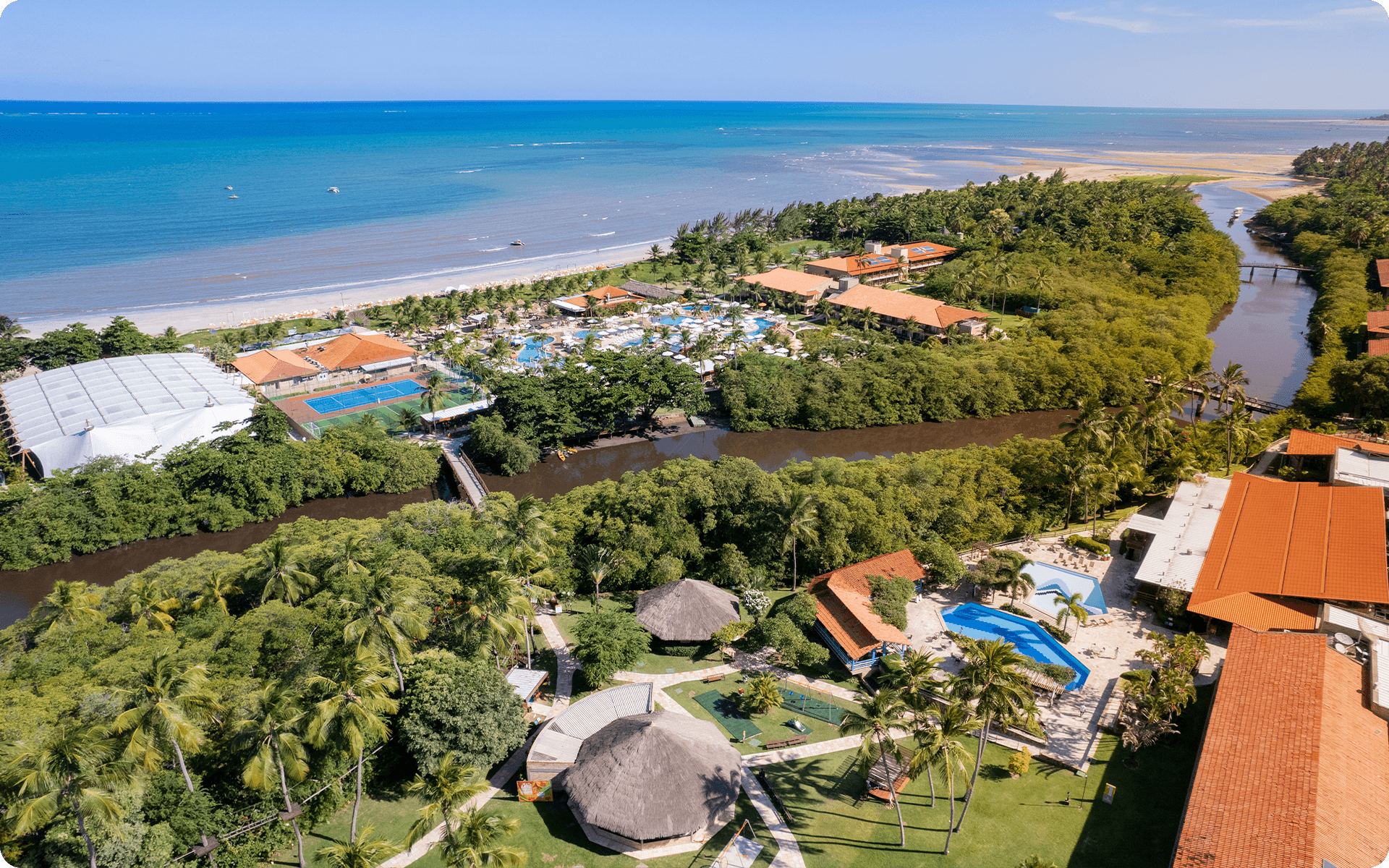 Vista do complexo do Salinas Maragogi, é possível ver as piscinas, pontos de consumo e o rio que cruza o resort. Ao fundo a Praia de Maragogi com maré baixa e dia ensolarado.