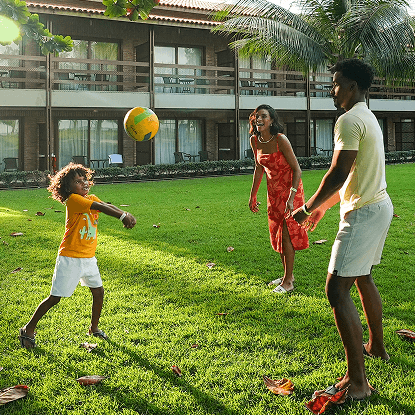 Família jogando volei na grama no fim de tarde, todos estão sorrindo. Estão em frente aos quartos em frente ao mar., o céu tem algumas nuvens.