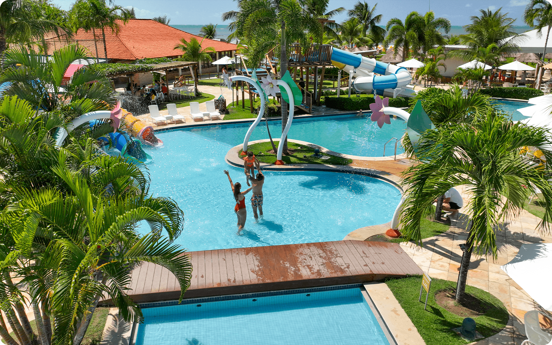 Em piscina infantil, família se diverte, pai carrega a filha no colo. Há uma ponte de madeira que cruza a piscina, e ao fundo tem os brinquedos infantis e muitos coqueiros.