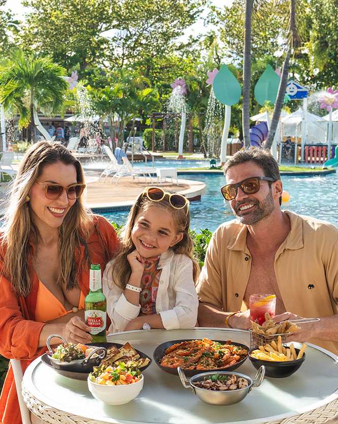 Família sorridente com pai, mãe e filha fazendo um lanche em uma mesa branca ao lado da piscina com brinquedos aquáticos infantis do resort Salinas Aaragogi. Sobre a mesa, há porções de camarão, batata frita e bolinhos.