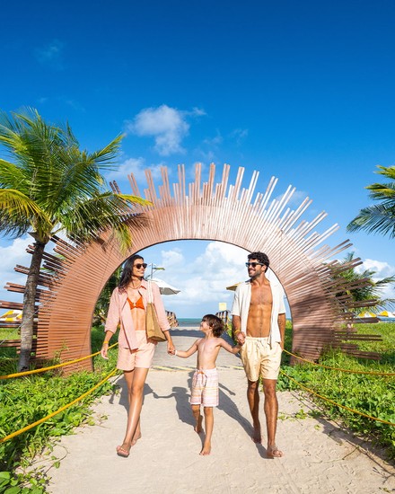 Férias em família no Salinas Maragogi All Inclusive Resort Família de três pessoas caminhando de mãos dadas em um caminho de areia que passa por baixo de um arco de madeira e dois coqueiros grandes, em direção ao Resort.