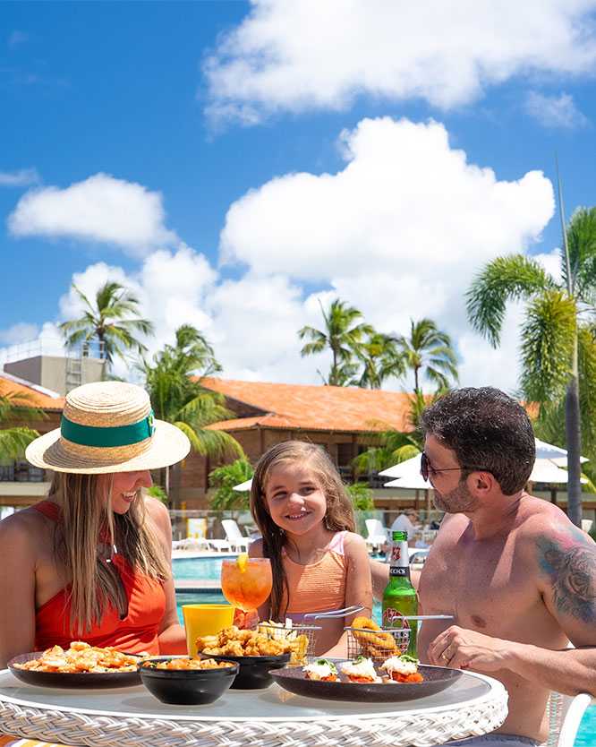 Família sorridente com pai, mãe e filha posando pra foto em uma mesa com diversos pratos de comida e bebidas, ao lado da área da piscina. Ao fundo, algumas espreguiçadeiras, guarda-sóis, coqueiros e a estrutura do Salinas Maragogi.