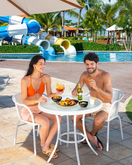 Resort All Inclusive em Maragogi com petiscos na Piscina Um casal sorridente sentado numa mesa sob um guarda-sol desfrutando de petiscos e bebidas, com a piscina e os escorregadores ao fundo.