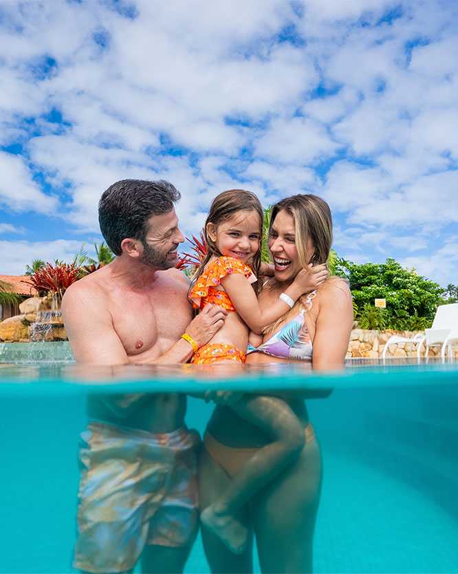 Família feliz, composta por pai, mãe e filha pequena, abraçados dentro da piscina azul do resort. A foto é tirada na altura da água, mostrando o céu azul com nuvens ao fundo, coqueiros e a arquitetura do Salinas Maragogi.