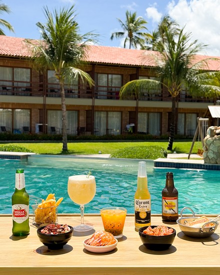 Aperitivos e Bebidas à beira da piscina do Salinas Maragogi Uma mesa de madeira com cervejas, drinks, batatas fritas e porções de frutos do mar, com a piscina e a fachada do resort ao fundo.