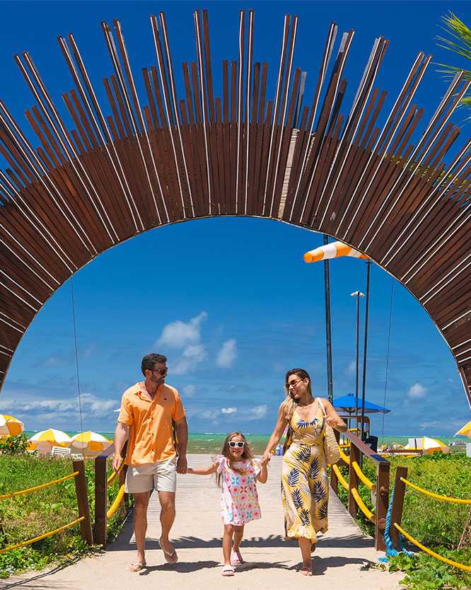 Família com pai, mãe e filha pequena de mãos dadas, caminhando sorridentes em direção à câmera, passando por dentro de um grande arco de madeira que serve de portal pra a praia do resort, com coqueiros e o céu azul ao fundo.