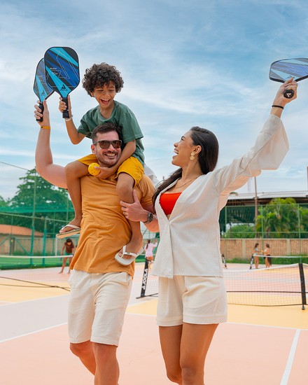 Salinas Maragogi: lazer, esportes e diversão em família Família sorridente com raquetes na mão celebra numa quadra de pickleball, com o filho nos ombros do pai.