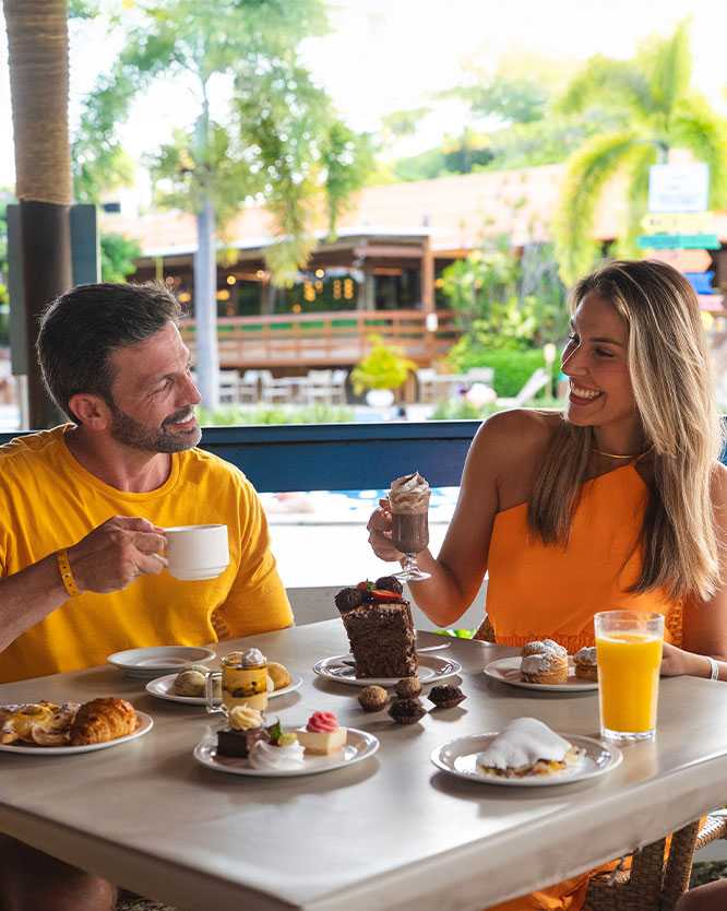 Família sorrindo e desfrutando de um lanche com muitos doces, salgados e bebidas em uma mesa. A mãe segura um copo com chantilly, o pai uma xícara e a filha ta na frente de um suco. A mesa está cheia de bolos, doces finos e salgados.