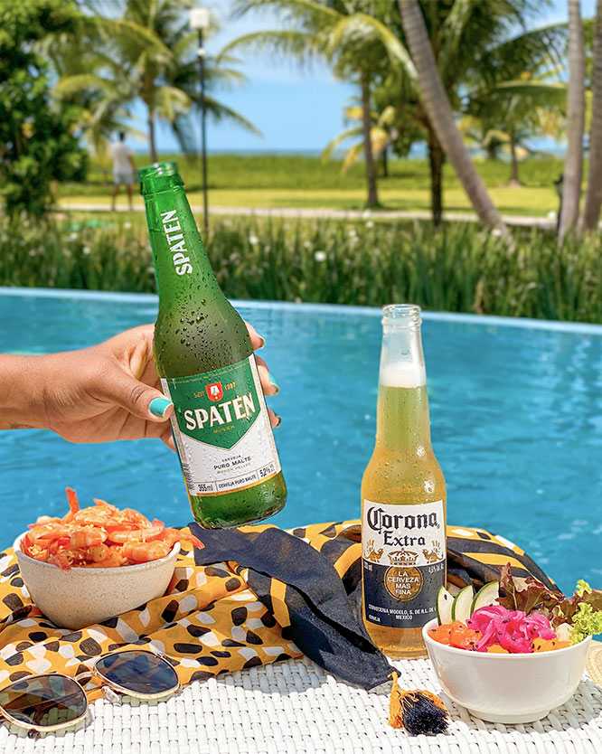 Mão segurando uma garrafa de cerveja Spaten. Sobre uma canga colorida na borda da piscina, estão uma garrafa de Corona Extra, óculos de sol e dois bowls de petiscos frescos. Ao fundo, coqueiros e o mar.