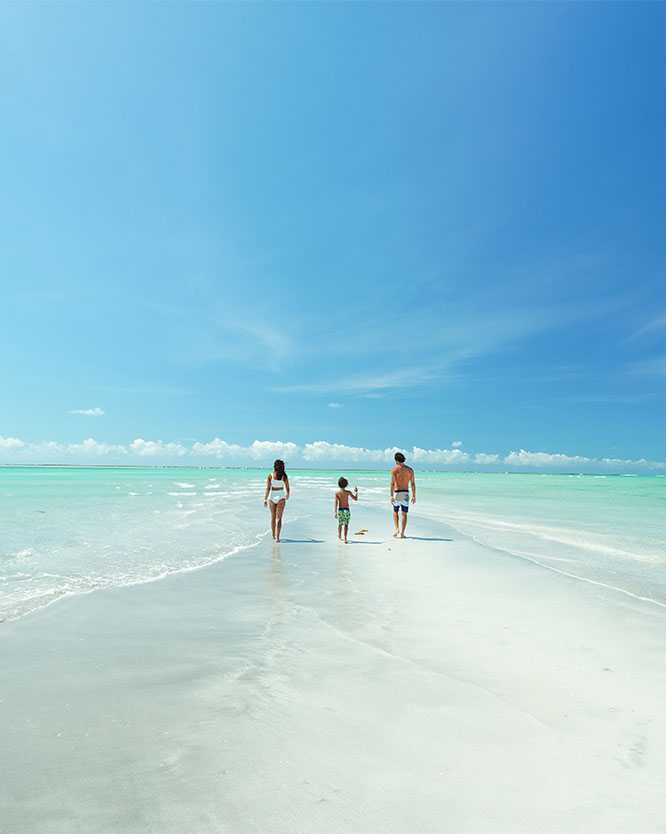 Fanília caminhando sobre faixa de areia branca que surge na maré baixa entre o mar azul turquesa da praia de Antunes, em Maragogi.