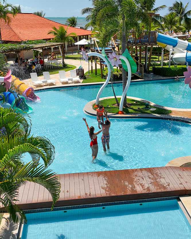 Família na piscina infantil do Salinas Maragogi. O pai segura a filha nos braços e a mãe está de braços abertos dentro da piscina para crianças do resort.