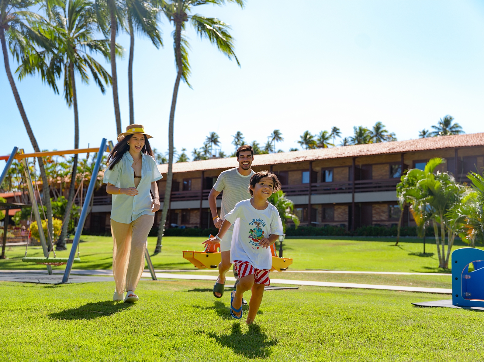 Família com pai, mãe e filho pequeno correndo e sorrindo no gramado do Salinas Maragogi, com palmeiras, parquinho e blocos de apartamentos ao fundo.