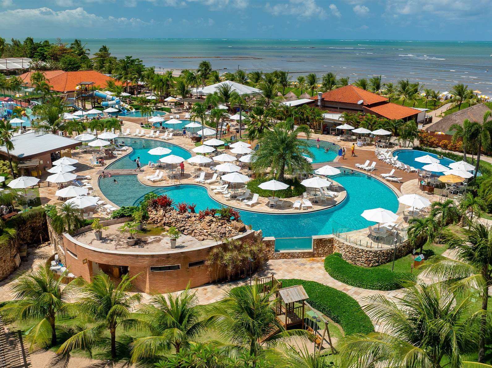 Vista aérea do Salinas Maragogi com complexo de piscinas cercadas por coqueiros, hóspedes curtindo as áreas de lazer, estruturas do resort e o mar ao fundo.