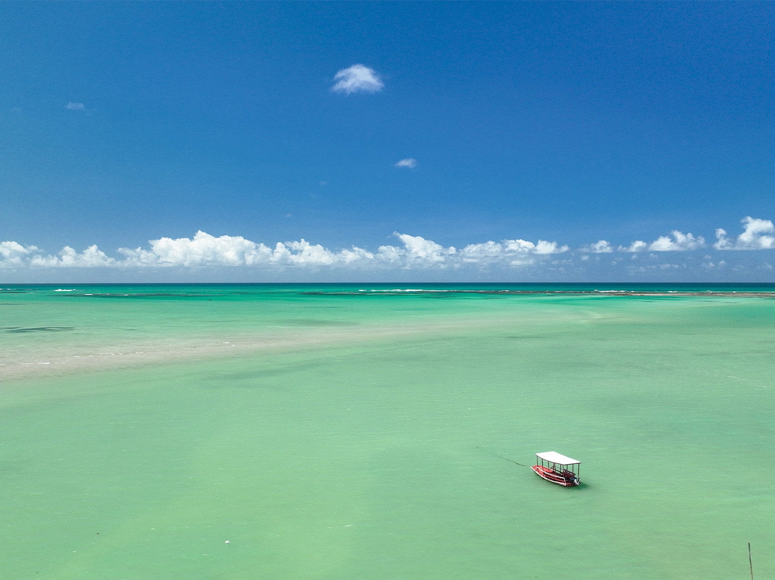 Vista aérea do mar em tons de verde cristalino da praia de Ponta de Mangue, com jangada navegando em águas calmas e céu azul com poucas nuvens ao fundo.