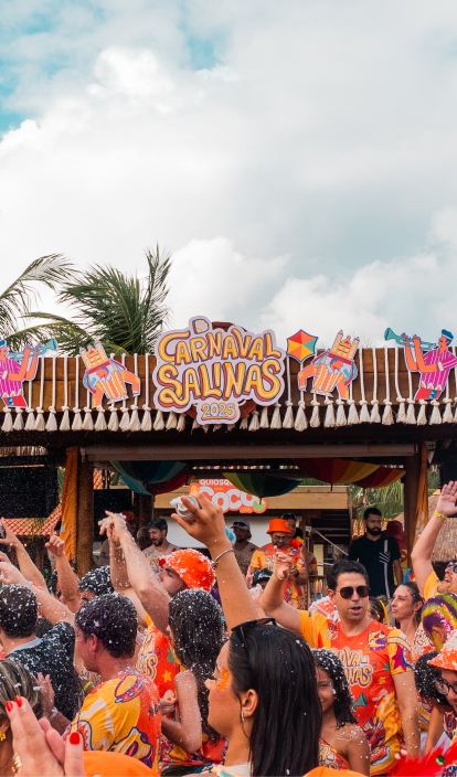 Bloco de carnaval no Salinas Maragogi, com pessoas fantasiadas e curtindo, braços levantados, espuma no ar e palco decorado com temática carnavalesca ao ar livre.