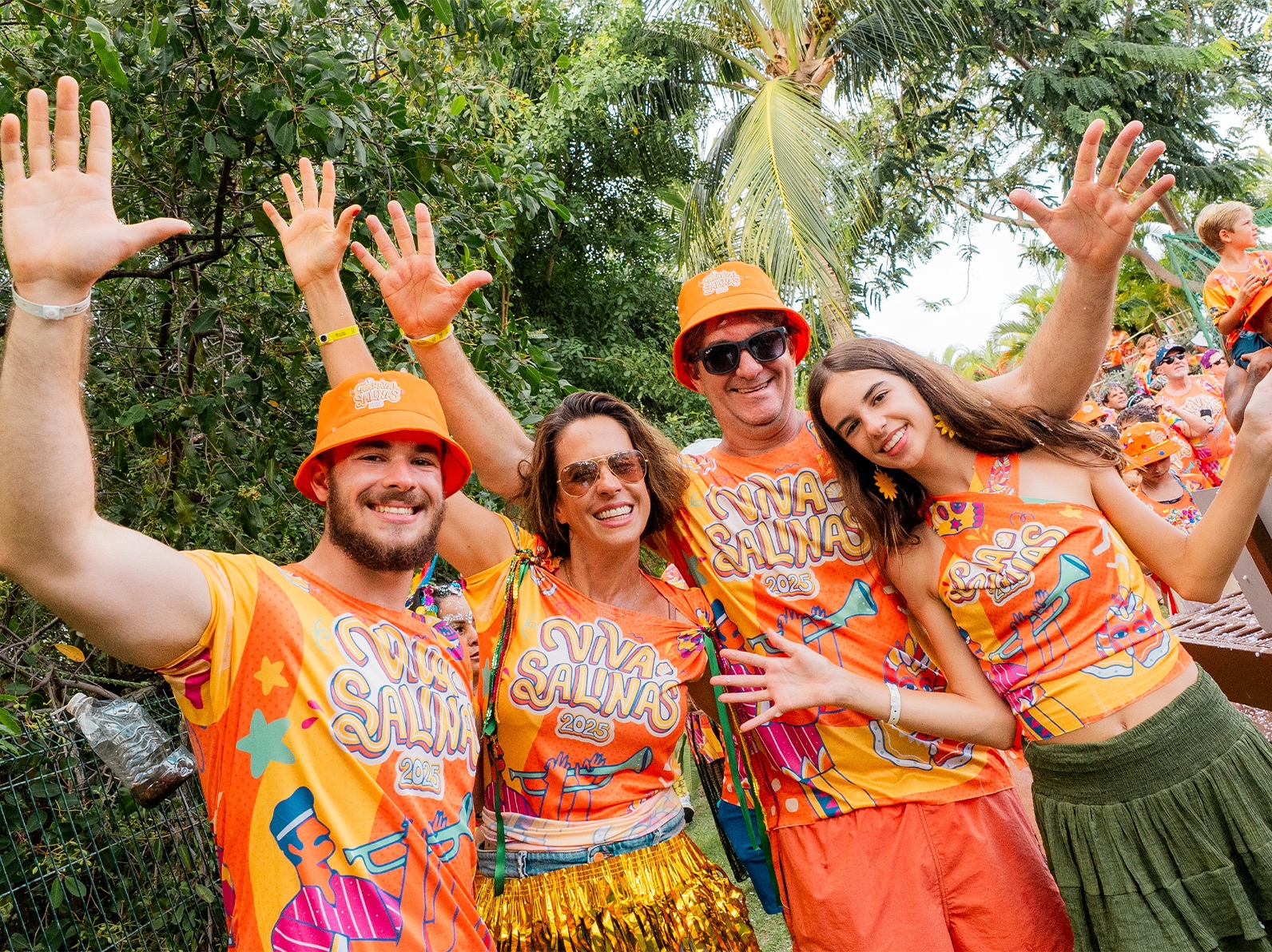 Família de quatro pessoas sorrindo e celebrando com os braços levantados, com abadás laranjas escritos Viva Salinas 2025 no resort Salinas Maragogi.