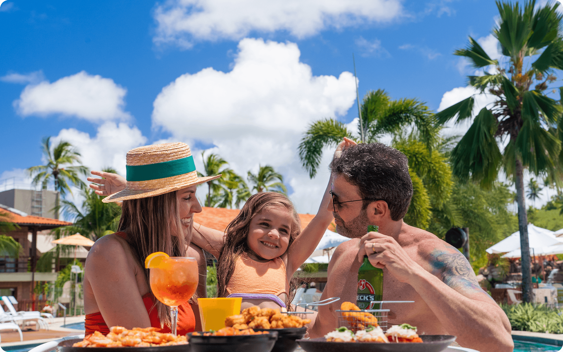 Família desguntando de petiscos e bebidas numa mesa, todos estão sorrindo, o dia é ensolarado e com poucas nuvens.