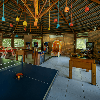 O teen space com repletos jogos disponíveis, mesa de pingue e pongue, sinuca, pebolim, cartas, fliperama, jogo de tabuleiro. No teto há pendentes de luz coloridos.