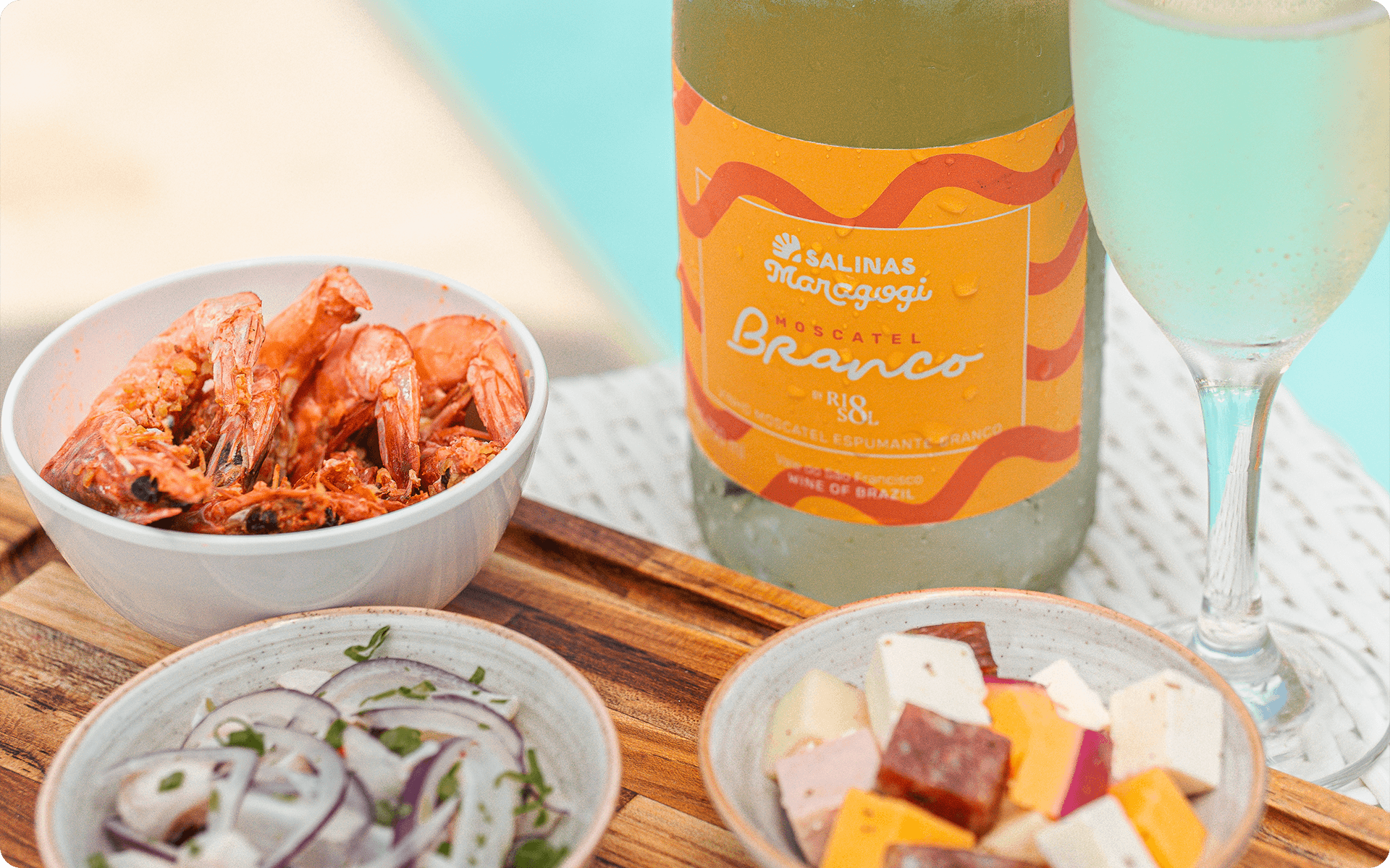 Moscatel branco do Salinas Maragogi com taça ao lado, e alguns petiscos como camarão, ceviche e queijos.