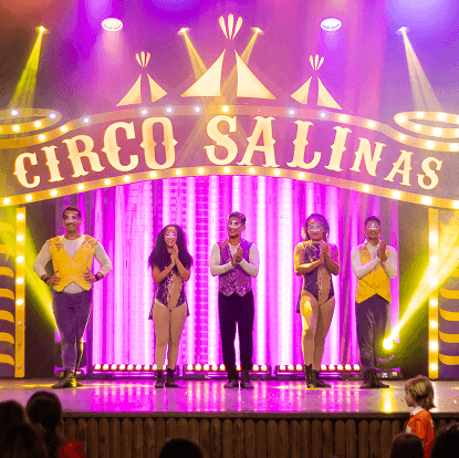 Personagens do Circo Salinas no palco do Espaço Cabanos se apresentando. Há luzes coloridas, o letreiro Circo Salinas ao fundo do palco.