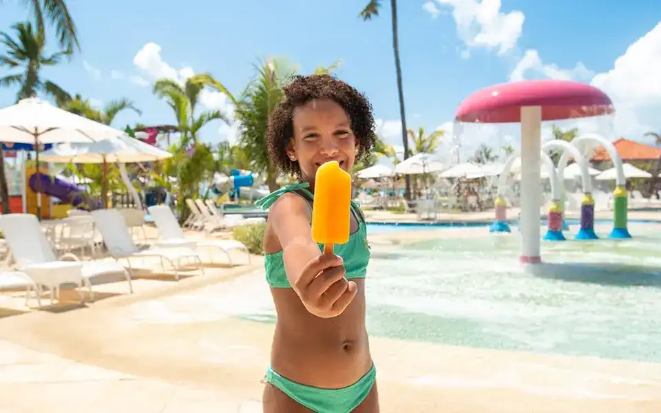 Uma criança sorridente, vestindo um biquíni verde, segura um picolé laranja com um grande sorriso no rosto. Ela está em primeiro plano, com um parque aquático infantil ao fundo.