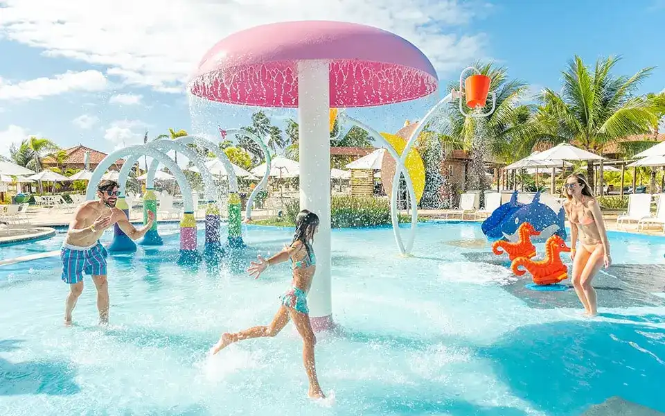 Uma criança com seus pais brinca sob um grande cogumelo  aquático e jatos de água coloridos. O céu azul claro e o sol brilhante realçam o ambiente tropical do resort, enquanto há palmeiras e a estrutura de lazer.