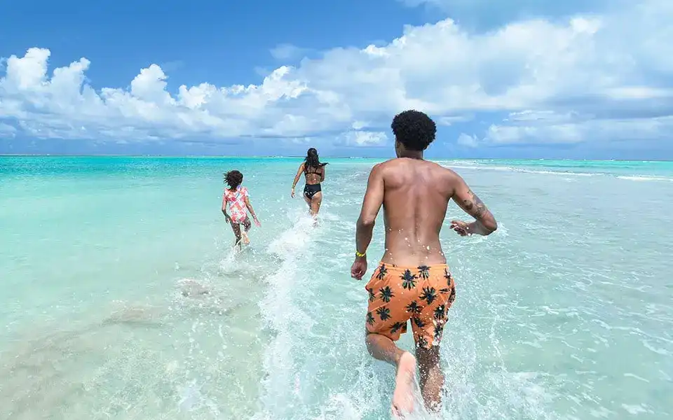 Três pessoas correm em direção ao mar cristalino sob um céu azul com nuvens brancas em Maragogi. A água transparente e rasa revela a areia abaixo, e a linha do horizonte ao fundo mostra um oceano calmo.