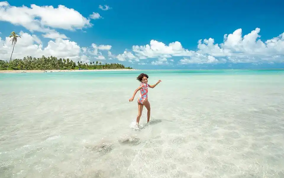 Uma criança feliz brinca na água rasa e cristalina de uma praia, com palmeiras e um céu azul pontilhado de nuvens brancas ao fundo. A menina usa um maiô colorido e sorri, transmitindo uma sensação de liberdade e prazer.