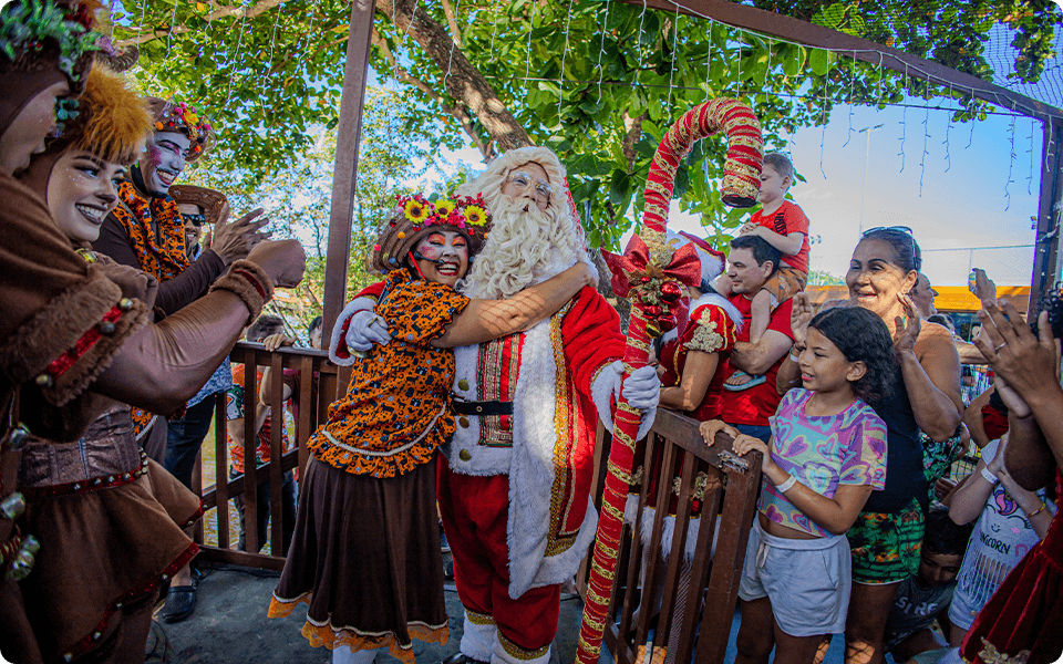 Papai Noel abraçando uma personagem e interagindo com famílias e crianças. Ao redor há muitas árvores.