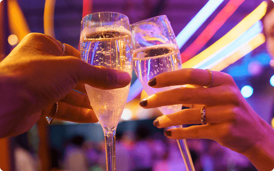 Duas taças de espumante sendo brindadas, com um fundo desfocado de festa com iluminação neon colorida (rosa, roxo, azul e amarelo).