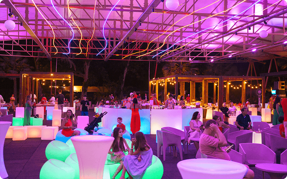 Área externa coberta com iluminação neon colorida, predominando tons de rosa e roxo, com famílias sentadas em pufes e cadeiras iluminados no Espaço Cabanos.
