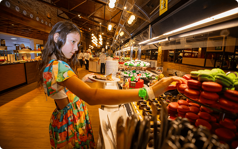 Criança em um buffet all inclusive, pegando doces decorados de natal em uma grande mesa de sobremesas, com cozinheiros ao fundo e ambiente interno com iluminação quente.