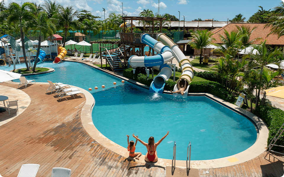 Piscina do resort Salinas Maragogi com toboáguas coloridos e uma mãe com sua filha na beira da piscina de mãos pro alto ao fundo tem a estrutura do Salinas Maragogi como campo, espreguiçadeiras.