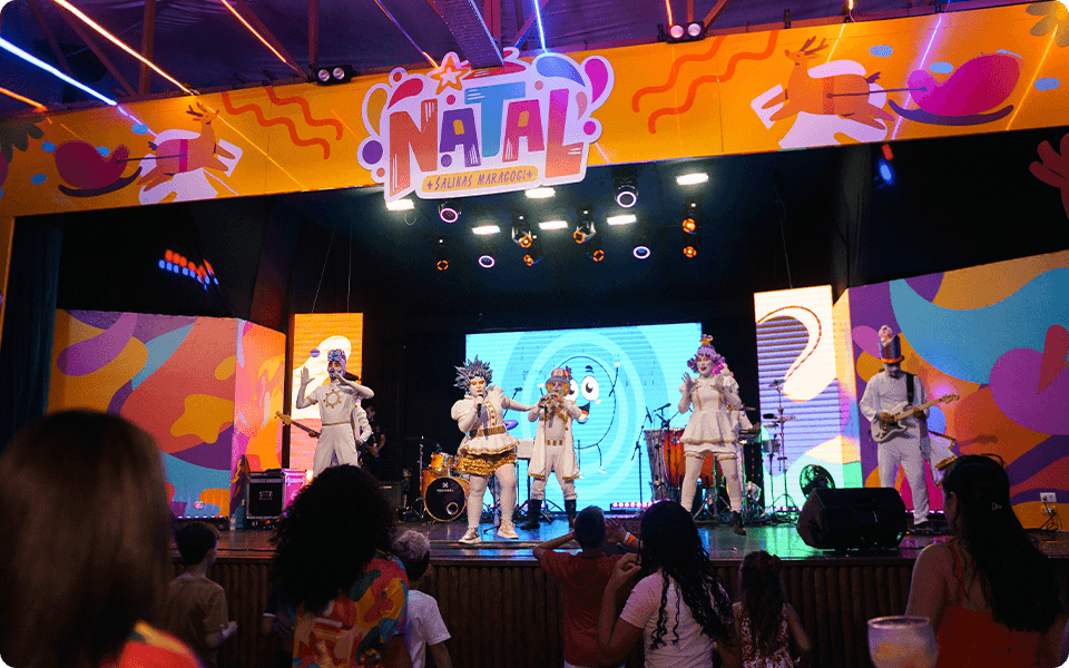 Show de Natal no palco do resort Salinas Maragogi, com banda, cantores fantasiados e famílias na plateia. Decoração natailina em todo o palco e um telão ao fundo.