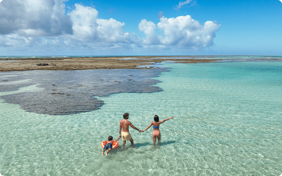 Família com uma criança em boia flutuando nas piscinas naturais de Ponta de Mangue, com águas cristalinas e céu com poucas nuvens.