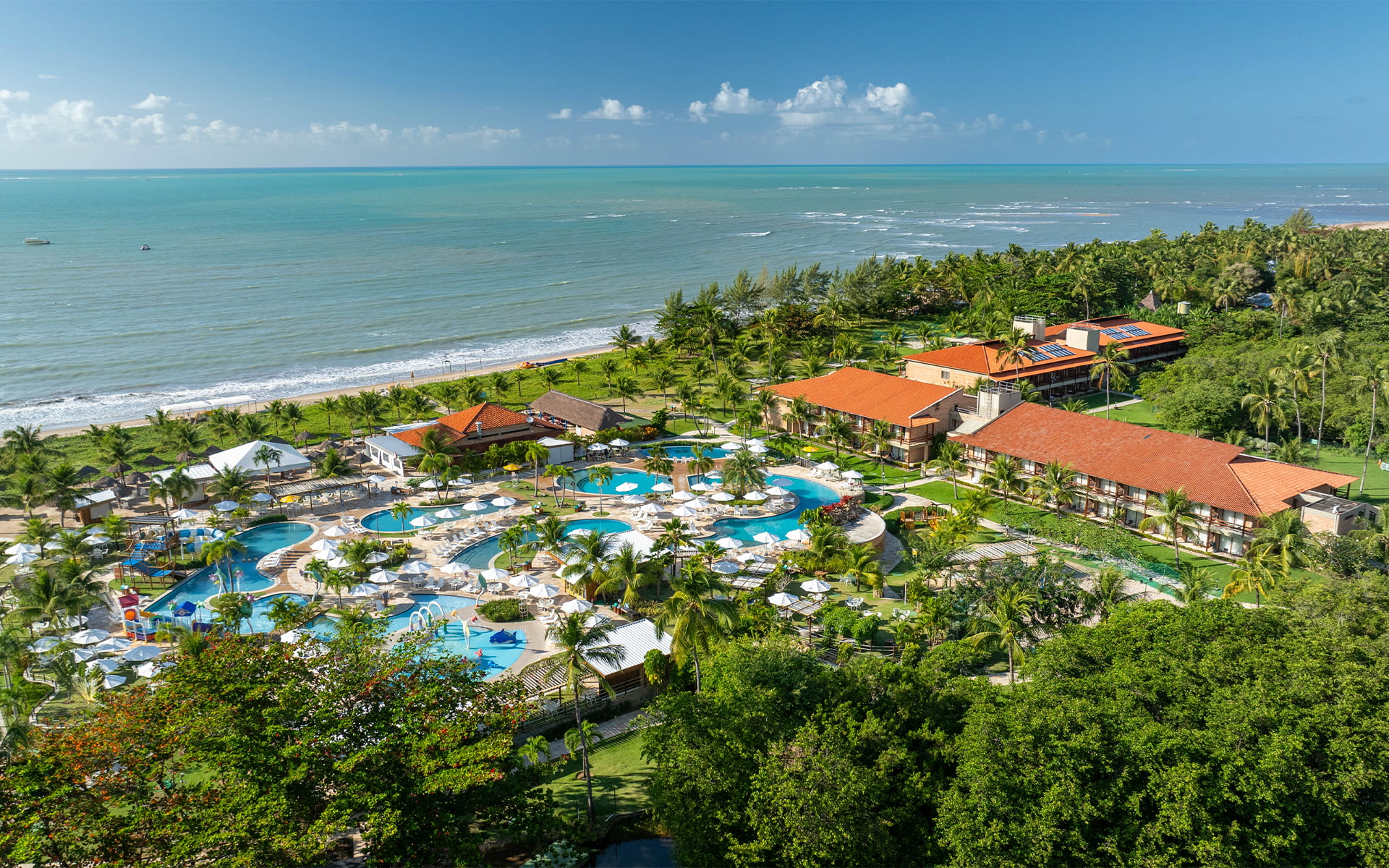 A estrutura completa do Salinas Maragogi All Inclusive Resort vista do alto. Vista aérea do Salinas Maragogi All Inclusive Resort apresentando o complexo de piscinas, parque aquático infantil, área de lazer com espreguiçadeiras e guarda-sóis, cercados por coqueiros e vegetação litorânea, além da praia ao fundo.