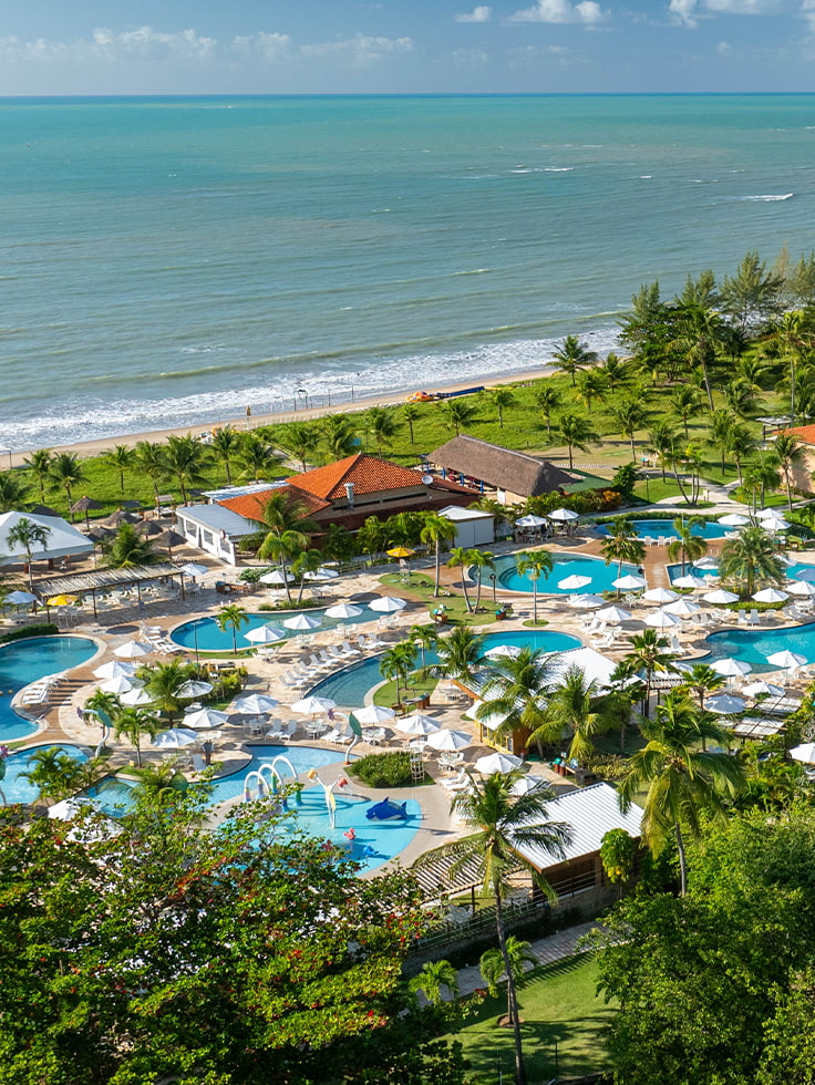 A estrutura completa do Salinas Maragogi All Inclusive Resort vista do alto. Vista aérea do Salinas Maragogi All Inclusive Resort apresentando o complexo de piscinas, parque aquático infantil, área de lazer com espreguiçadeiras e guarda-sóis, cercados por coqueiros e vegetação litorânea, além da praia ao fundo.