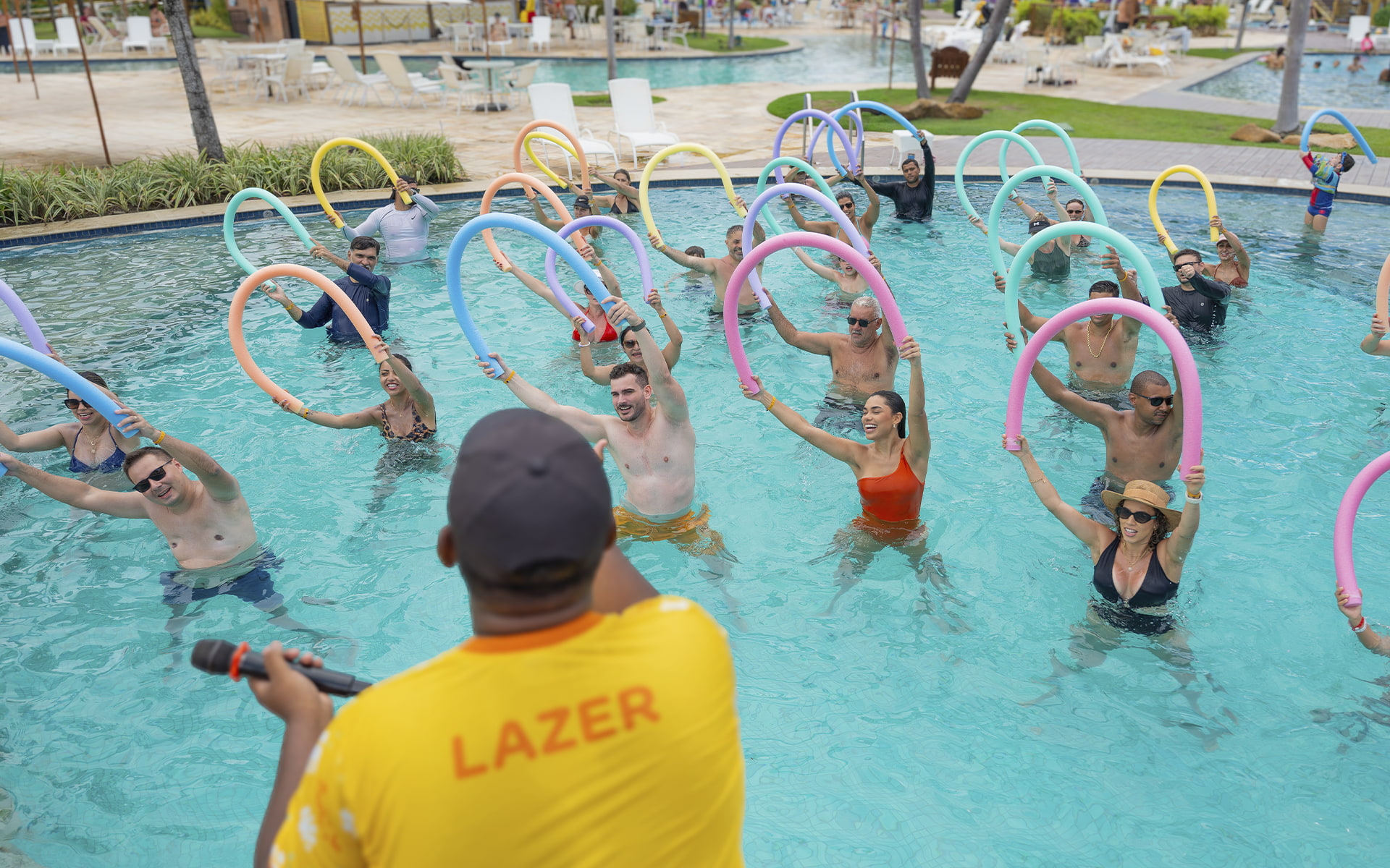 Experiências que contagiam e unem toda a família Monitor de lazer do Salinas Maragogi conduzindo aula de hidroginástica com espaguetes coloridos para grupo de hóspedes na piscina, todos com os braços erguidos e sorrindo.