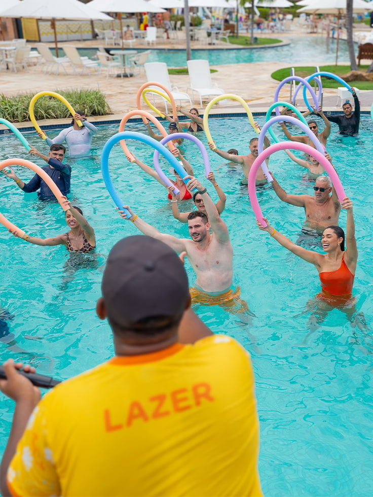 Experiências que contagiam e unem toda a família Monitor de lazer do Salinas Maragogi conduzindo aula de hidroginástica com espaguetes coloridos para grupo de hóspedes na piscina, todos com os braços erguidos e sorrindo.
