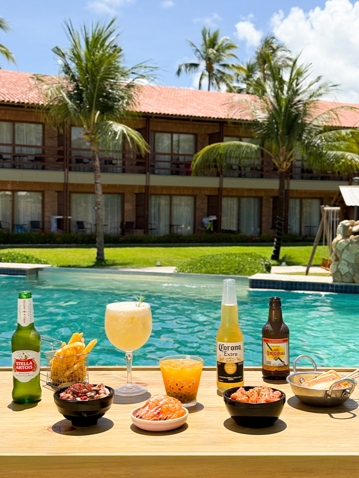 Sabores que combinam com o seu verão Mesa posta à beira da piscina do Salinas Maragogi com cervejas, drinks, batata frita, camarão e petiscos, com a estrutura dos quartos do Salinas Maragogi ao fundo.