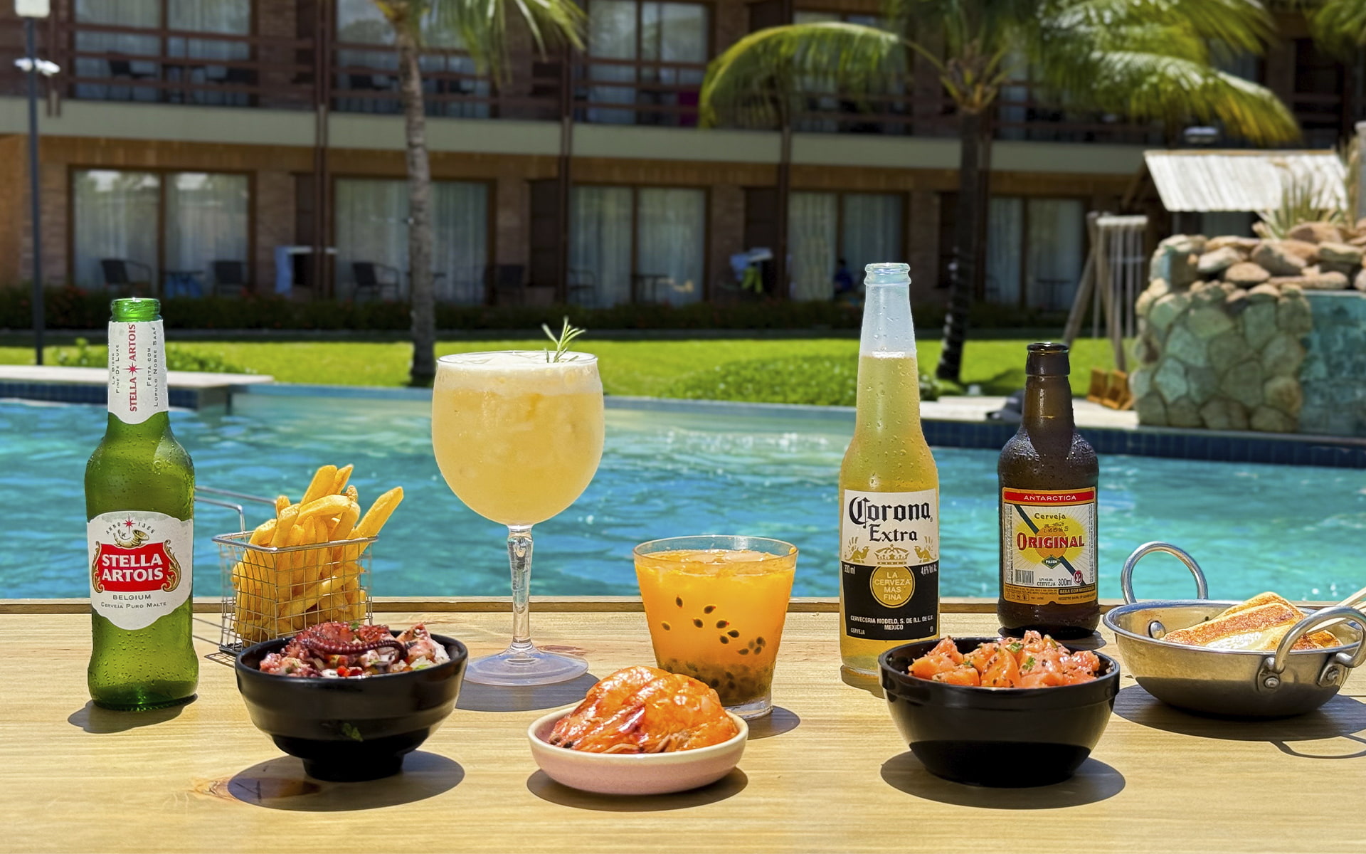 Sabores que combinam com o seu verão Mesa posta à beira da piscina do Salinas Maragogi com cervejas, drinks, batata frita, camarão e petiscos, com a estrutura dos quartos do Salinas Maragogi ao fundo.