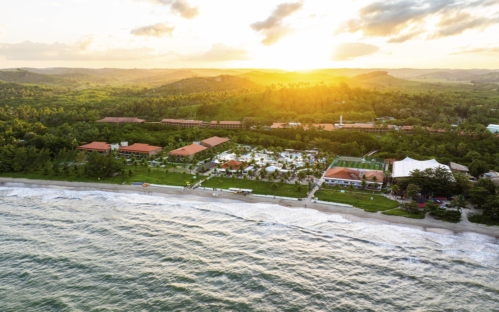 O pôr do sol que fecha o dia com chave de ouro Vista aérea do Salinas Maragogi ao entardecer, com o complexo de piscinas, praia e mata nativa ao fundo, sob céu dourado de pôr do sol.