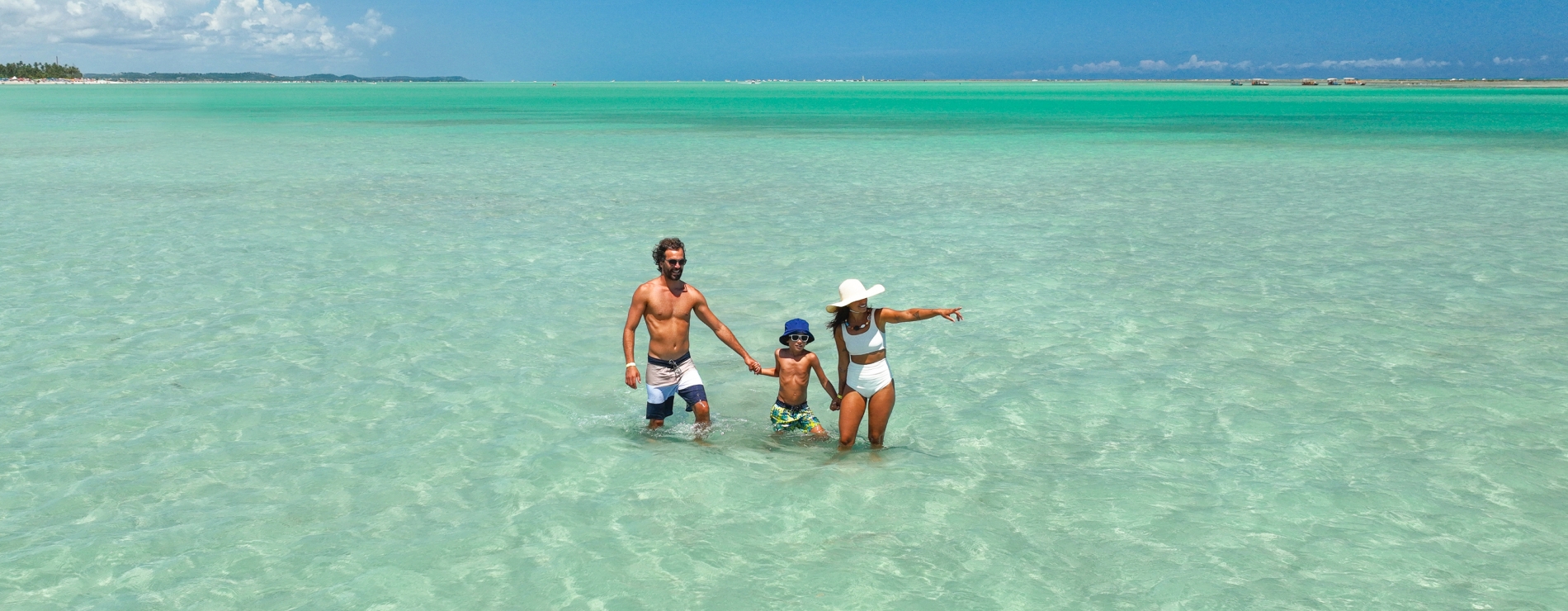 As águas cristalinas da Praia de Xaréu Família com pai, mãe e filho de mãos dadas caminhando nas águas rasas e cristalinas da Praia de Xaréu em Maragogi, com vegetação litorânea ao fundo em dia de céu azul.