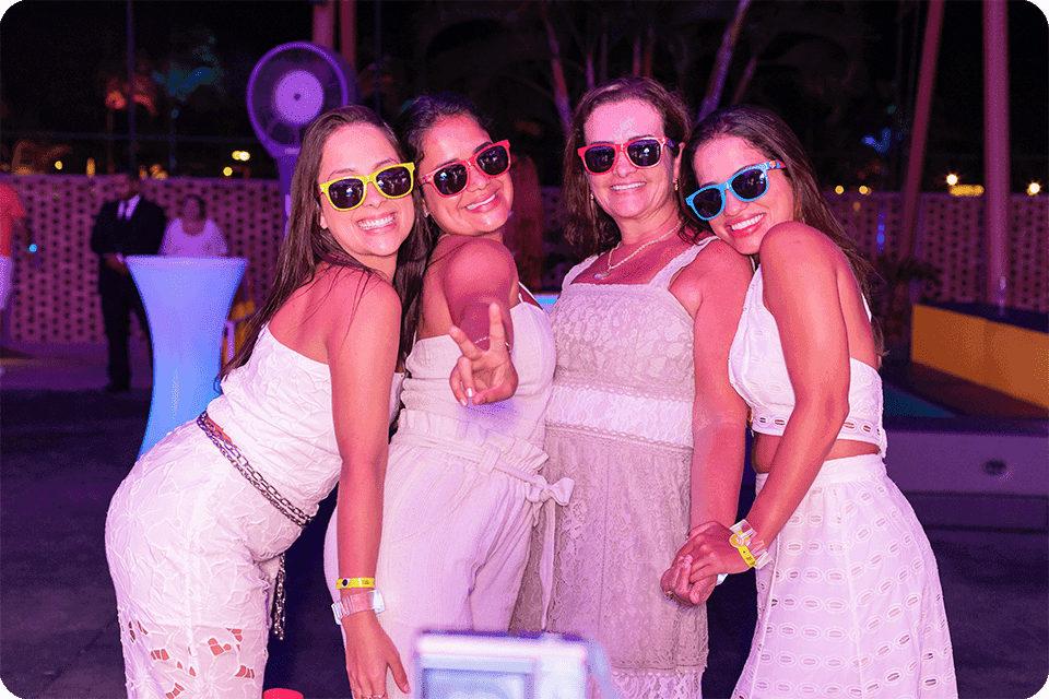  Quatro mulheres usando roupas brancas em comemoração ao ano novo e
 óculos escuros coloridos posam pra a foto, sorrindo, na festa do
 Réveillon no Salinas Maragogi.o