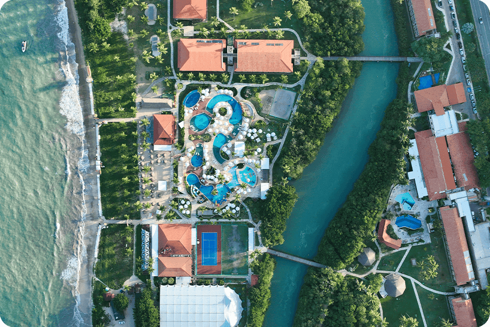 Vista aérea do Salinas Maragogi All Inclusive Resort, mostrando
a área das piscinas e algumas acomodações que são cortadas pelo Rio Maragogi. Todo o resort é cercado por vegetação tropical, palmeiras e está à beira-mar.