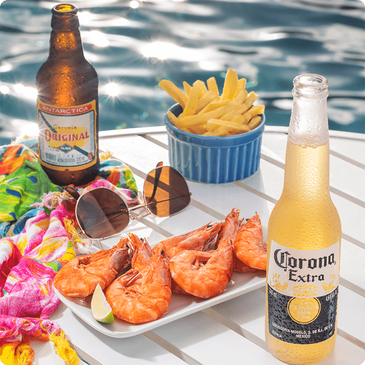 Cervejas Corona e Original, ao lado camarões e bata fritas como petiscos sobre mesa com óculos de sol e uma saída de praia colorida à beira da piscina no Salinas Maragogi.