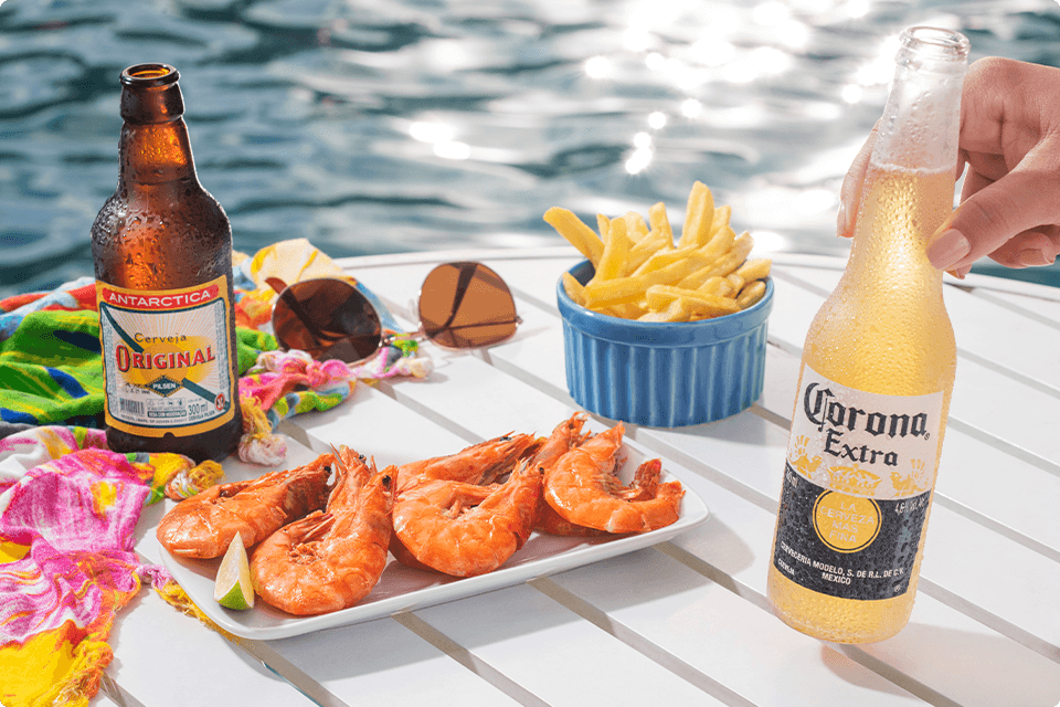 Cervejas Corona e Original, ao lado camarões e bata fritas como petiscos sobre mesa com óculos de sol e uma saída de praia colorida à beira da piscina no Salinas Maragogi.