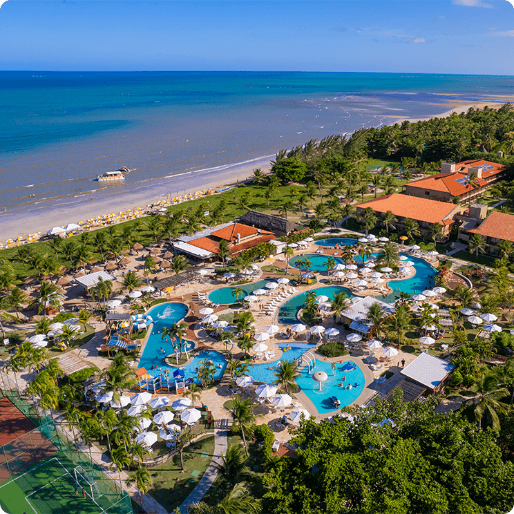 Vista aérea do Salinas Maragogi All Inclusive Resort, mostrando
a área de piscinas, guarda-sóis brancos e estrutura do resort, cercado por vegetação e está à beira-mar.