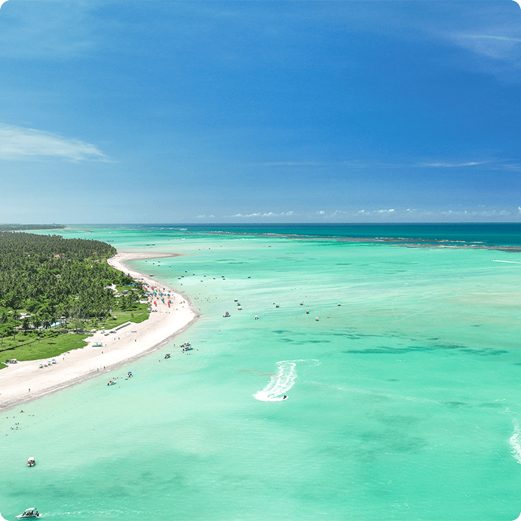 Vista aérea da Praia de Antunes com dia ensolarado e sem nenhuma nuvem. O mar está azul turquesa e bem raso podendo visualizar as piscinas naturais. Tem alguns barcos navegando no mar e pessoas andando e se divertindo na areia da praia, e tem uma grande vegetação de coqueirais ao fundo.