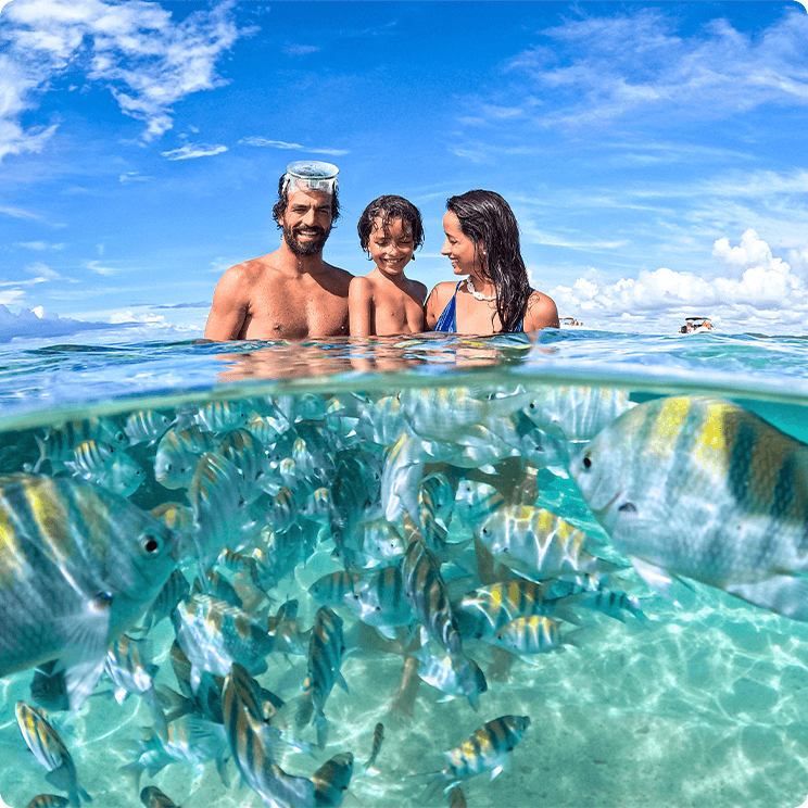 Foto subaquática com família sorridentes. Pai está com óculos de mergulho na cabeça e tem inúmeros peixe listados ao redor deles, o céu está com poucas nuvens.